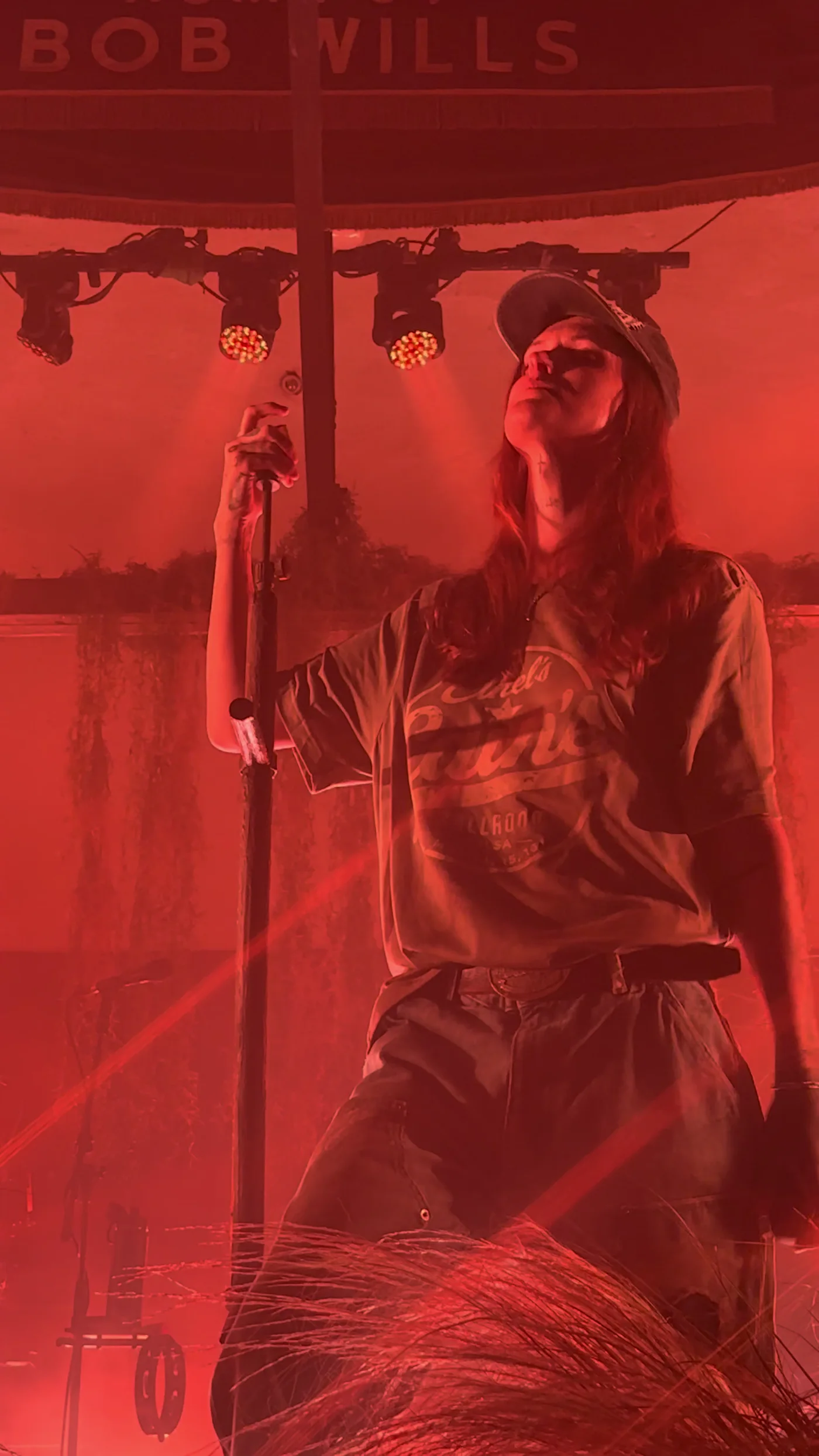 Ethel Cain lit hard red under the old Bob Wills sign, one hand on the microphone stand, head tipped back. Tulsa, Oklahoma · the last night of the tour. Frame from footage by @tempest.tossed, © the uploader.