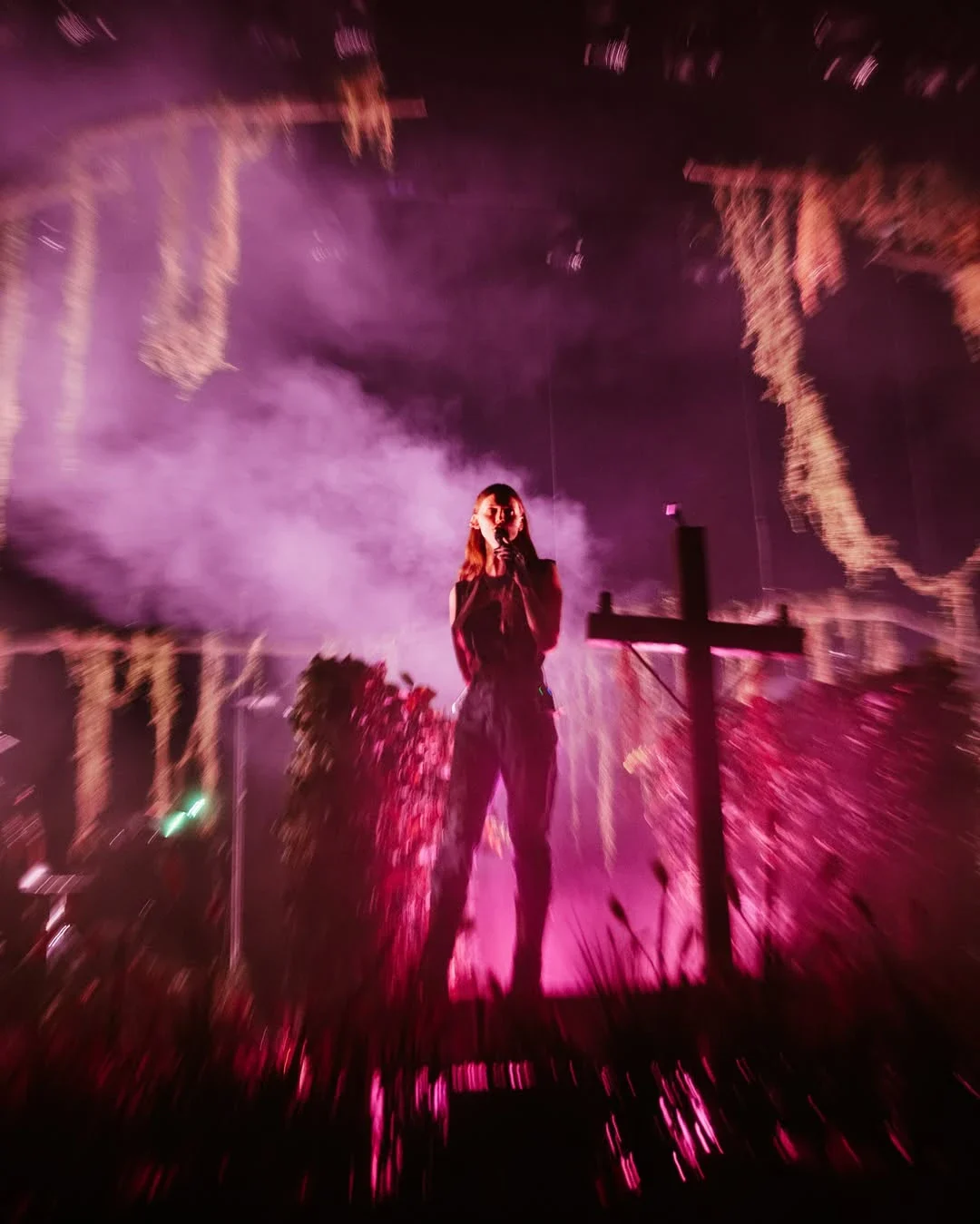 Ethel Cain alone at the centre of the stage in a column of pink smoke, the wooden cross beside her, moss hanging over the whole set. Paramount Theatre, Seattle · 12 August 2025, opening night. Photograph by an unnamed photographer, © the photographer.