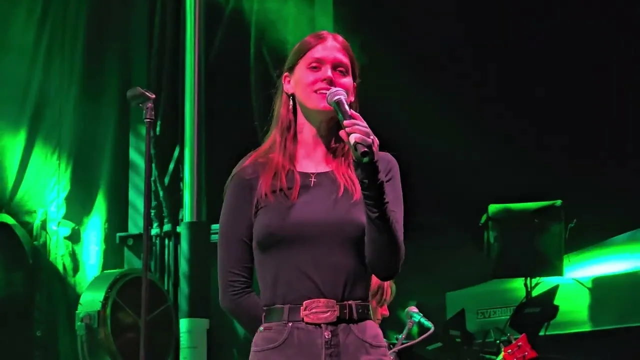 Ethel Cain close on a handheld microphone, the stage lit green behind her. Abiquiú, New Mexico · 14 September 2024. Frame from footage by Neroli Laine, © the uploader.