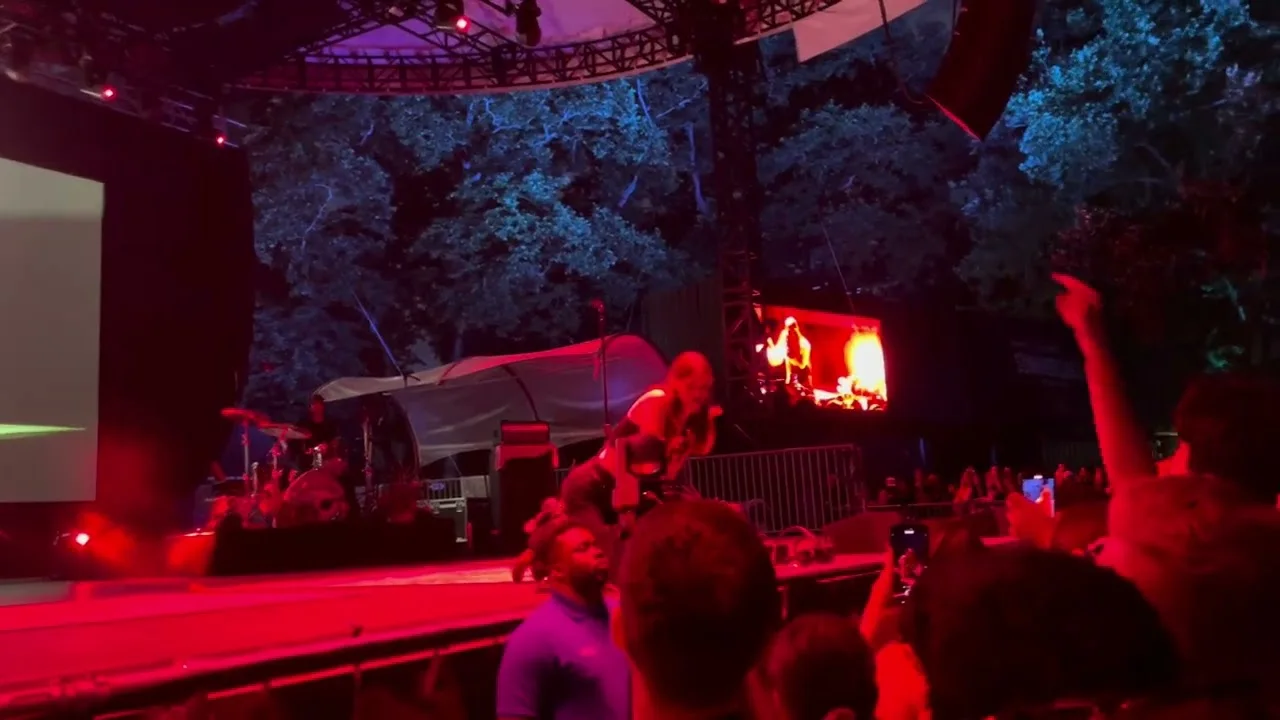 Ethel Cain small and red-lit at the far end of the stage, the crowd a dark mass under the trees. Central Park SummerStage, New York · June 2024. Frame from footage by Luna Monroe, © the uploader.