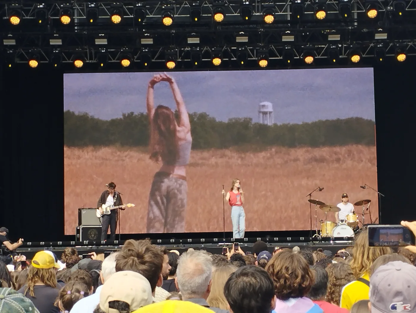 Ethel Cain in a red vest on a wide festival stage, the screen behind her filled with a dry field and a distant water tower, the crowd in silhouette. August 2023. Photograph by ManoSolo13241324, CC BY-SA 4.0.