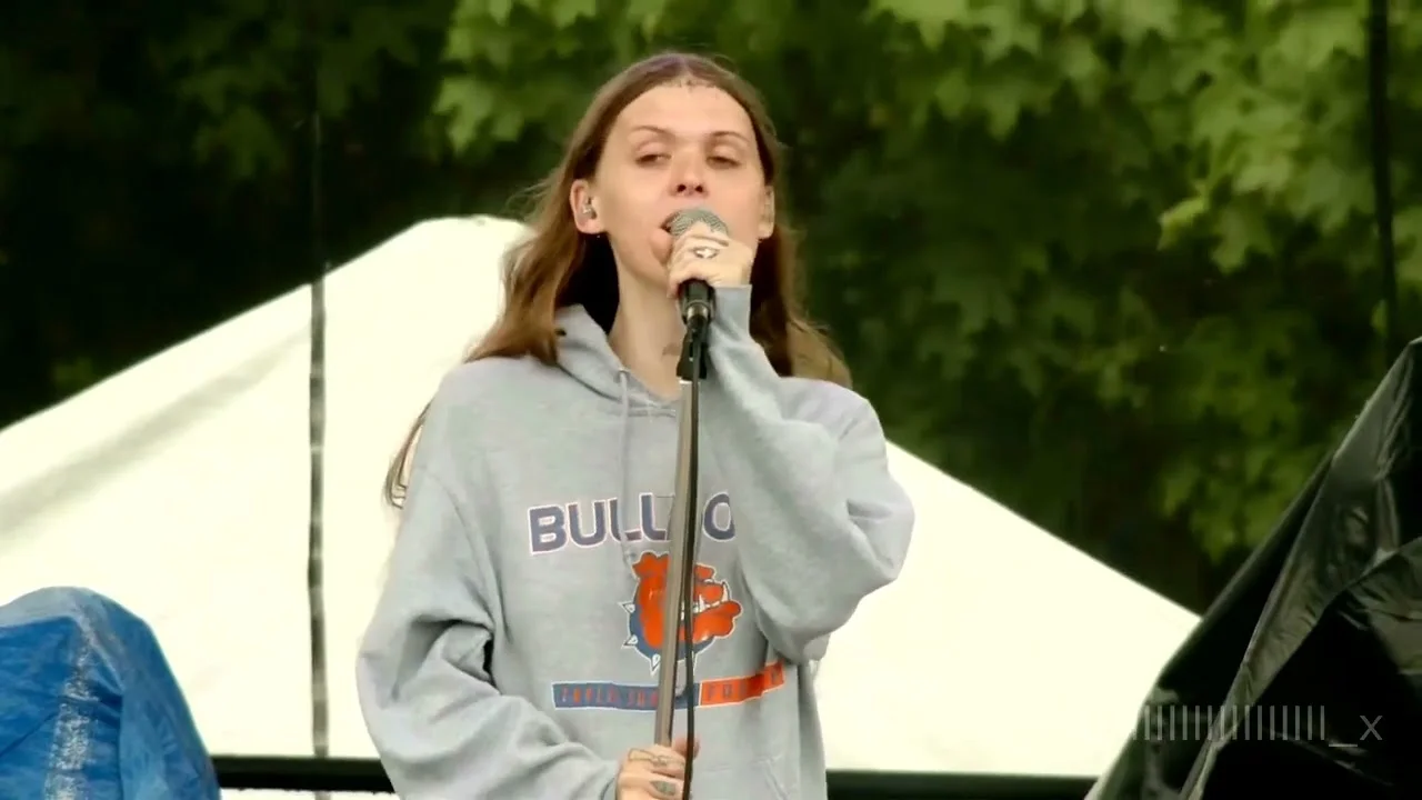 Ethel Cain singing into a mic stand in daylight, in an oversized grey hoodie, the white peak of a festival tent behind her. Pitchfork Music Festival, Chicago · July 2022. Frame from footage by Andrew Cronin, © the uploader.
